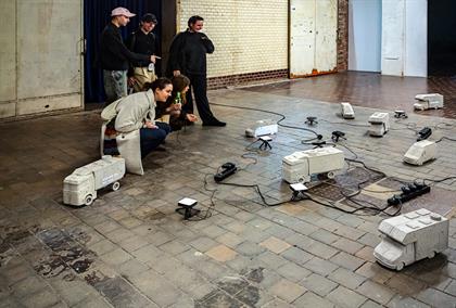 Zu sehen sind einige Studierende die auf ein Modellkonstrukt aus verschiedenen Miniatur Camper und ein Netz aus Beleuchtungen schauen, in einer Lagerhalle.